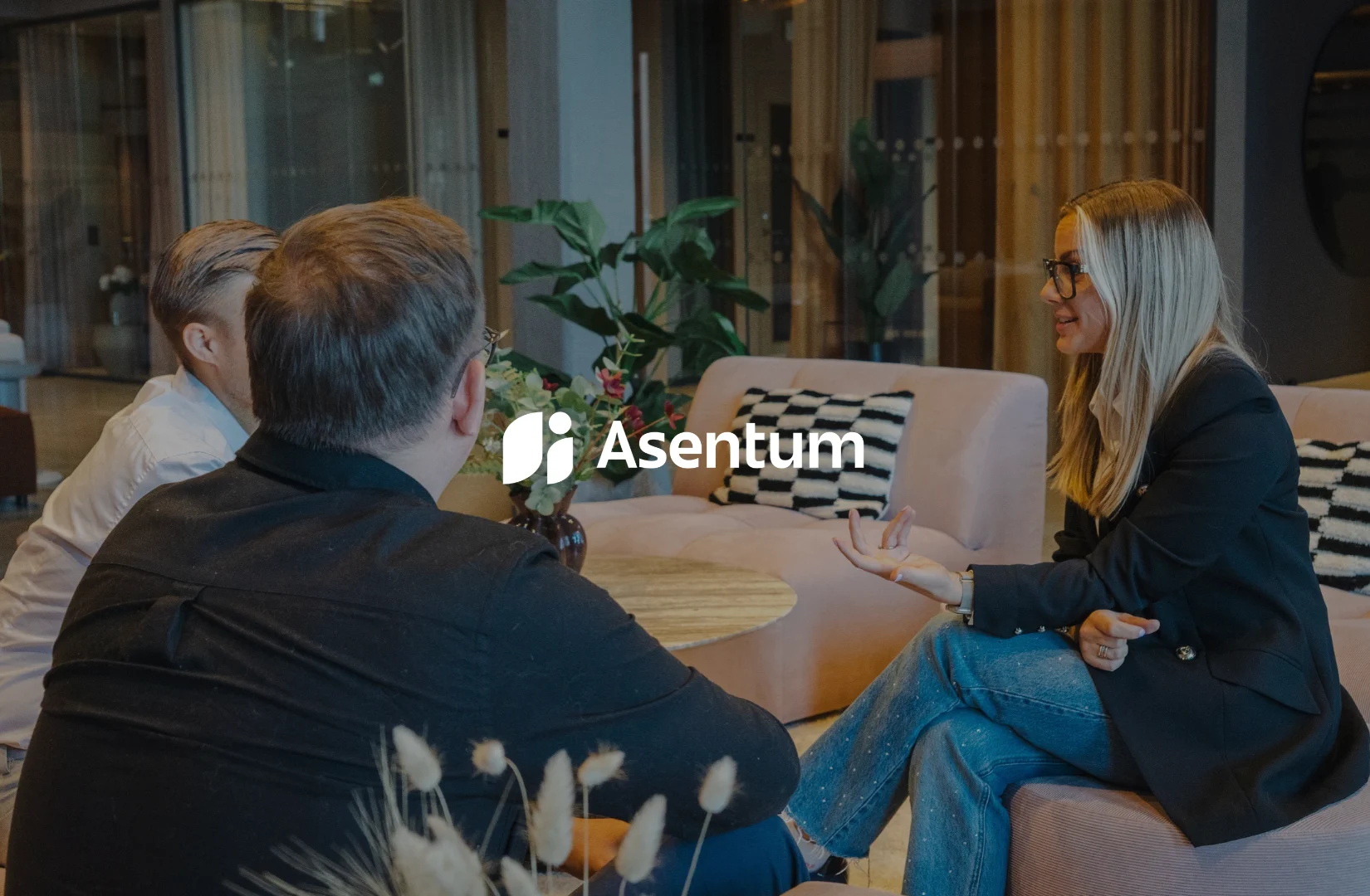Kvinna i glasögon samtalar med två män i en loungemiljö med Asentums logotyp över bilden