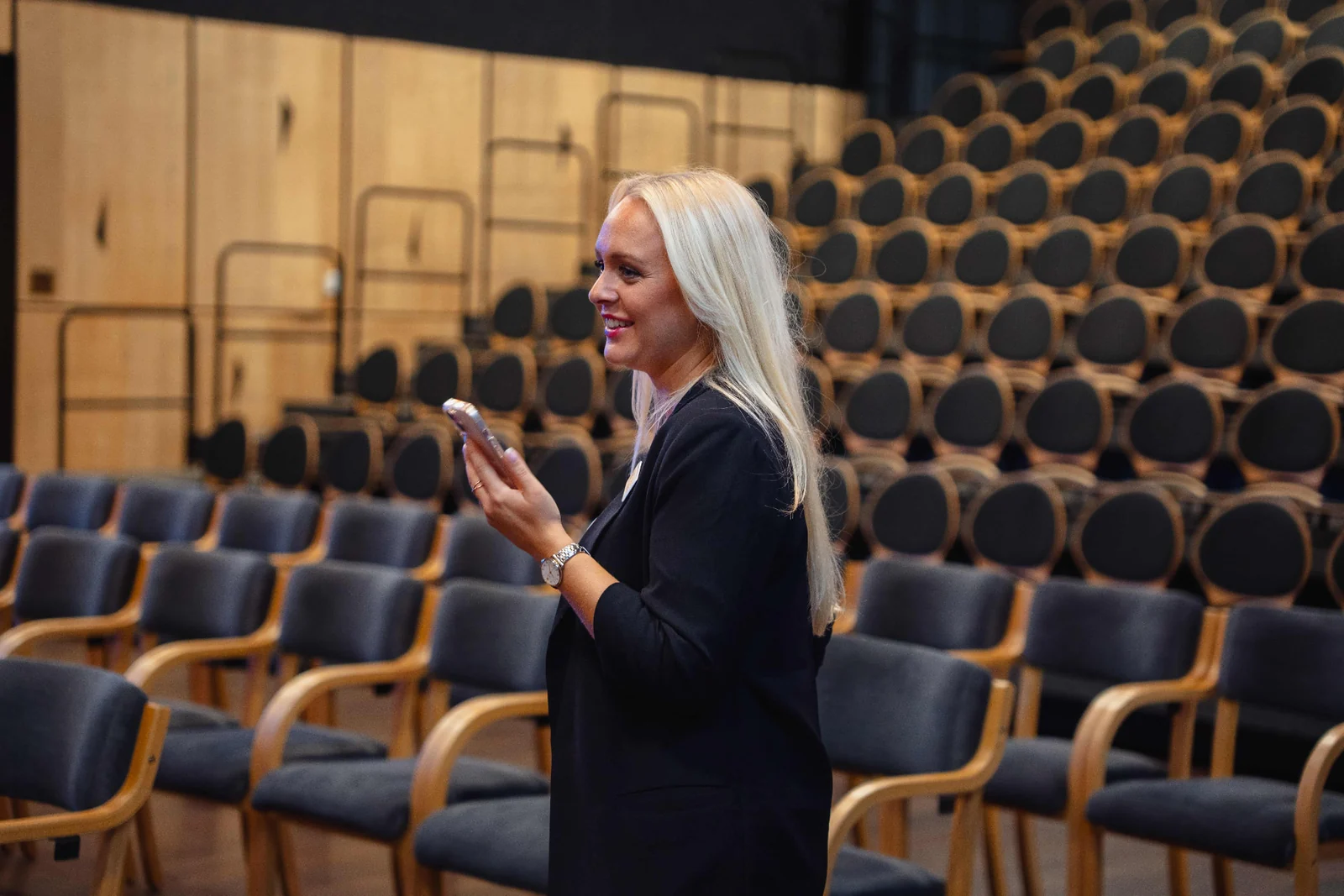 Kvinna i svart kavaj med mobiltelefon, leende framför rader av tomma stolar i ett auditorium